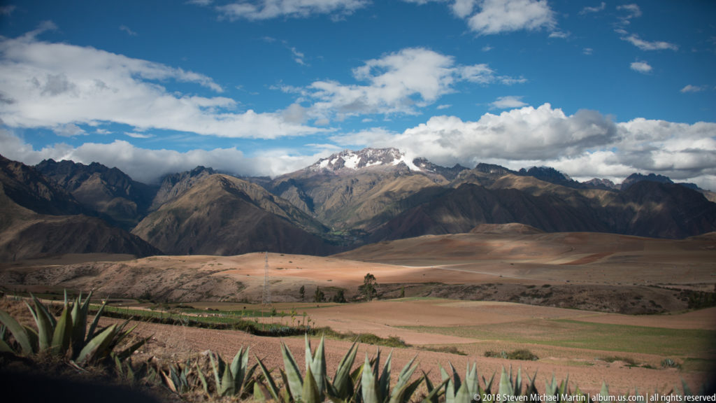 Cultivated Andean fields and agave-like plants below rugged mountains with snow patches and broken clouds.