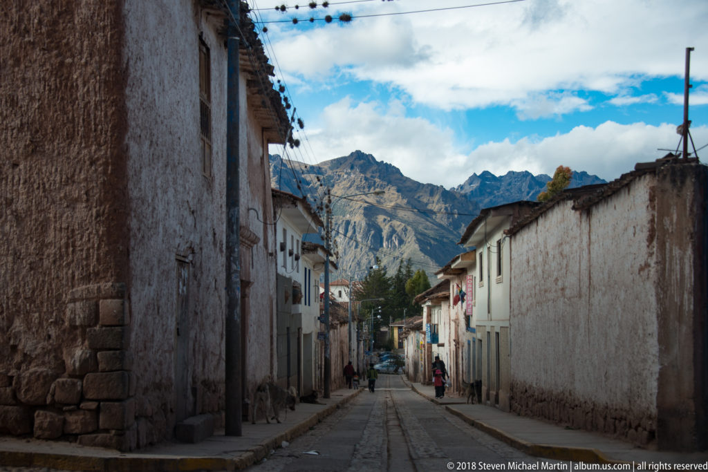 Narrow village street between whitewashed buildings, with pedestrians and jagged Andean mountains visible beyond.