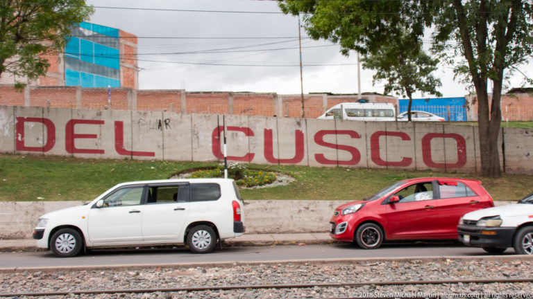 Large red DEL CUSCO lettering on a weathered wall behind parked vehicles, with railway tracks in the foreground.