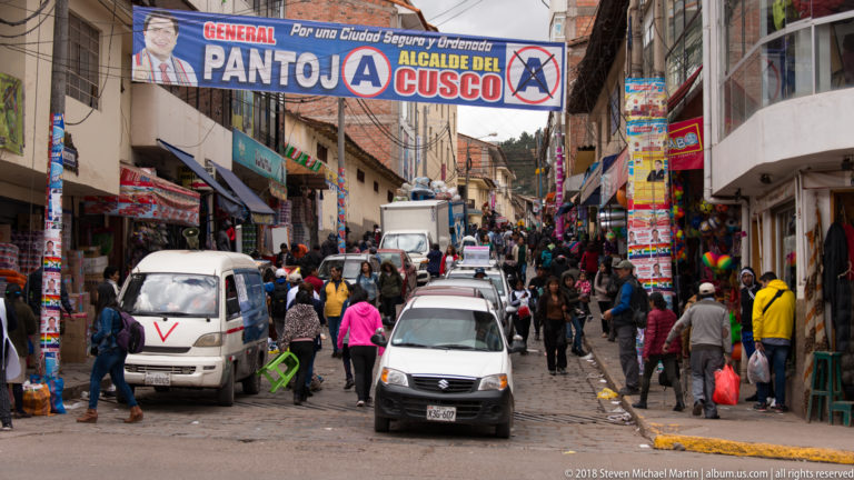 Congested commercial street with cars, vans, pedestrians, shops, and an overhead Alcalde del Cusco campaign banner.