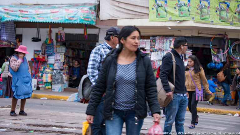 Woman carrying shopping bags crossing a commercial street in front of densely stocked shops and other pedestrians.