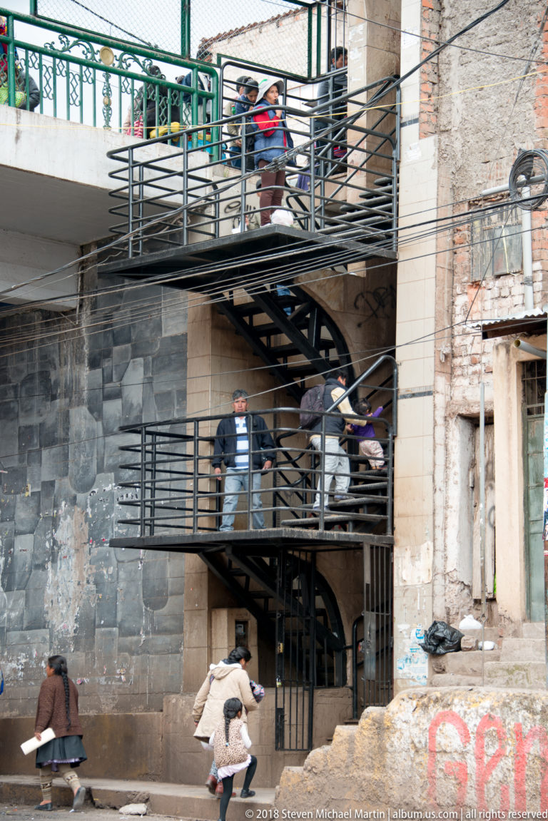 Exterior metal stairs and stacked balconies on a masonry building, with people on upper levels and pedestrians below.