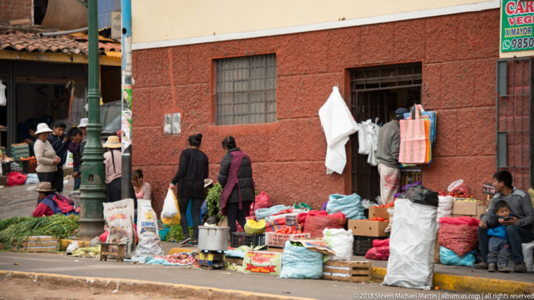 Curbside market along a red wall with leafy vegetables, sacks, crates, dry goods, vendors, and customers.