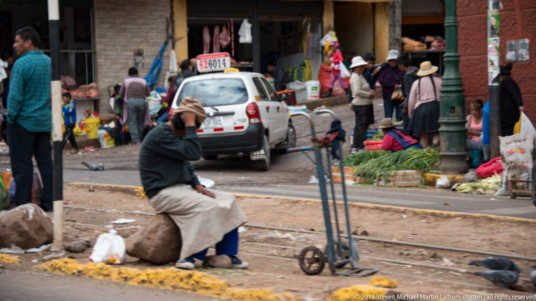 Seated person beside railway tracks facing a busy produce market, parked white car, pedestrians, and hand truck.