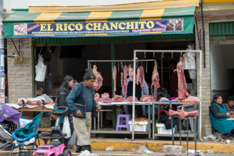 Open-front butcher shop with large raw meat cuts hanging from hooks and displayed on tables as pedestrians pass.
