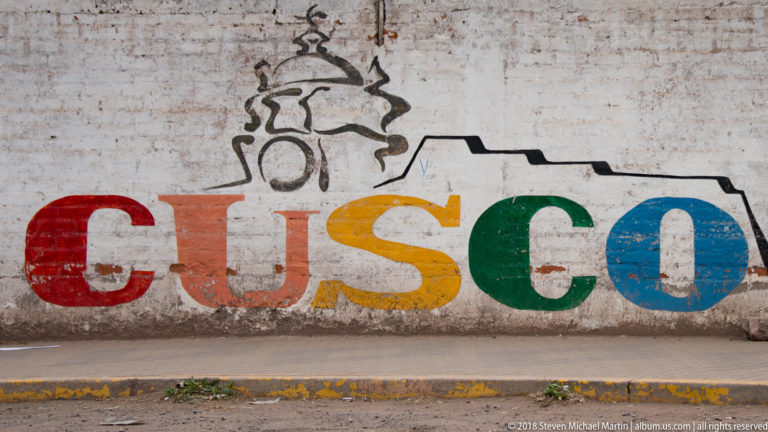 Large multicolored CUSCO letters painted on a weathered white wall beneath a black stylized line drawing.