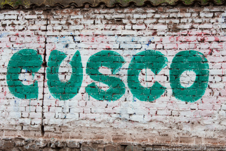 Large green CUSCO lettering painted across a weathered white brick wall with older paint showing through.