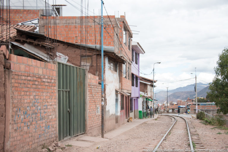 Curving railway tracks running beside unfinished brick houses and narrow paths, with mountains in the distance.
