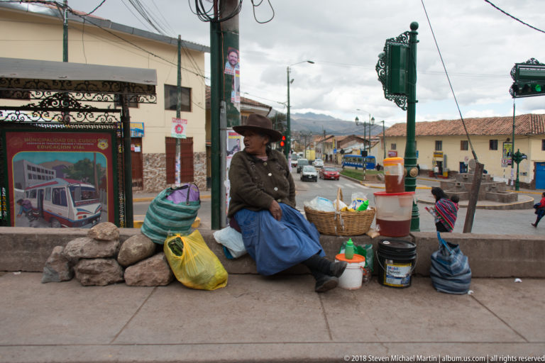 Seated woman in a brown hat and blue skirt surrounded by baskets, sacks, and buckets at a street intersection.