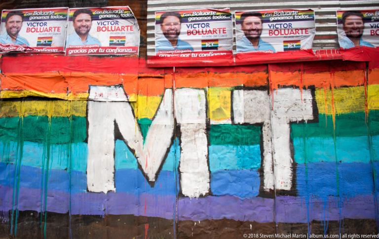 Large white MT initials over rainbow stripes beneath repeated Víctor Boluarte campaign posters.