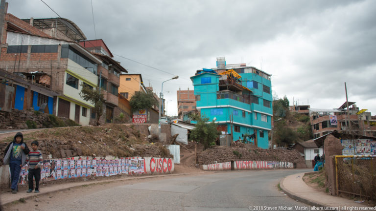 Curving highland road between brick buildings and a turquoise structure, with pedestrians and campaign-painted walls.