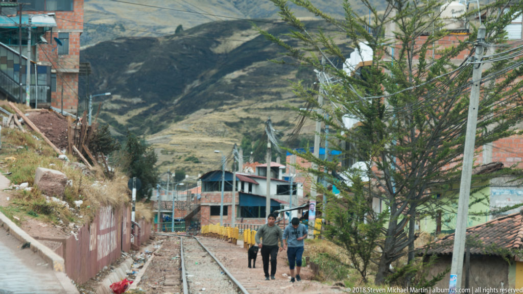 Three pedestrians walking along railway tracks between houses, with steep Andean mountains rising in the distance.