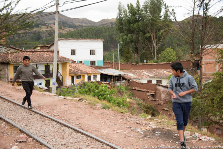 Two young men walking beside railway tracks among tile-roofed houses and unfinished brick buildings.