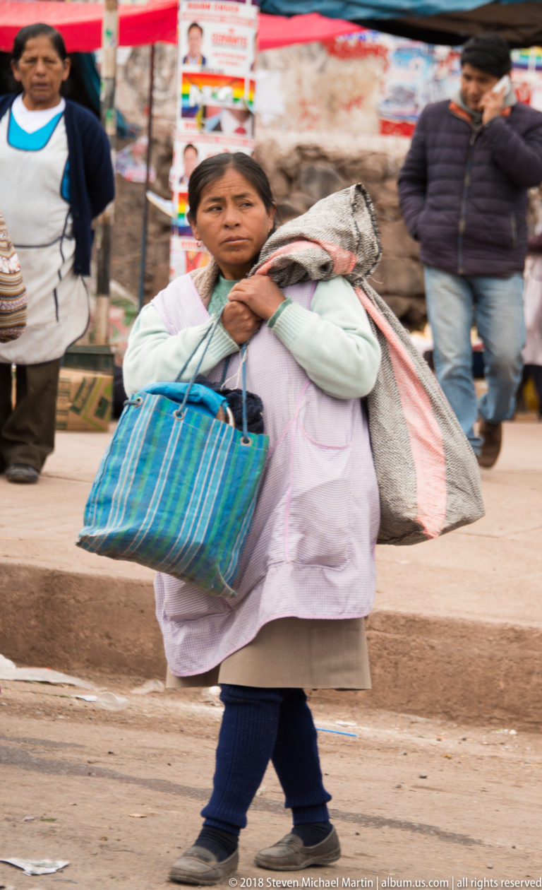 Woman walking through a market carrying a large striped bag and bulky sack over her shoulder.