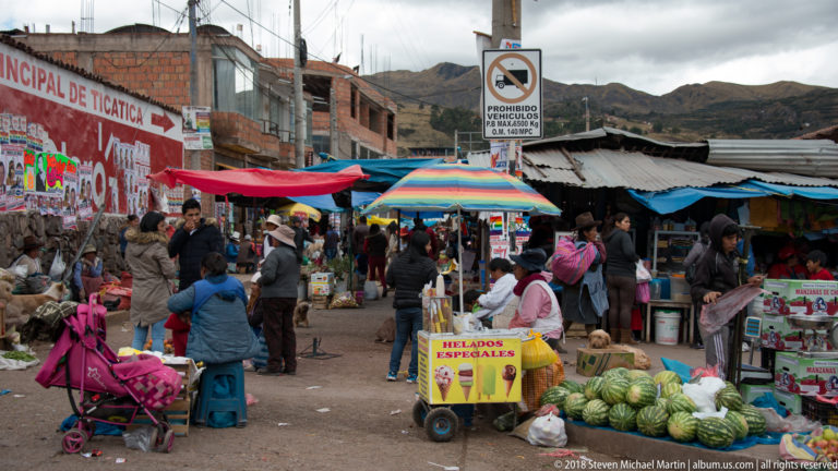 Busy open-air market lane with shoppers, produce, colorful umbrellas, watermelons, and a Helados Especiales cart.