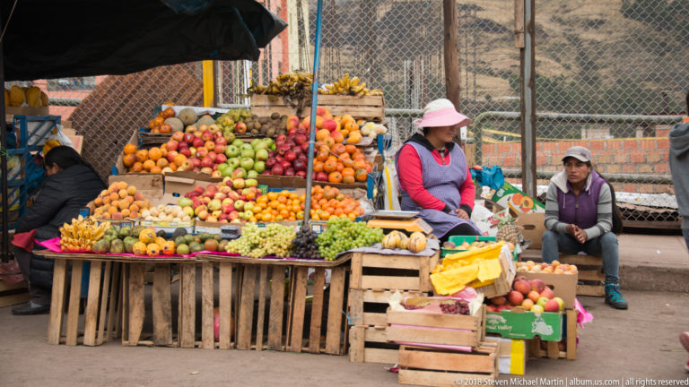 Open-air fruit stall piled with citrus, apples, bananas, grapes, and other produce around two seated vendors.