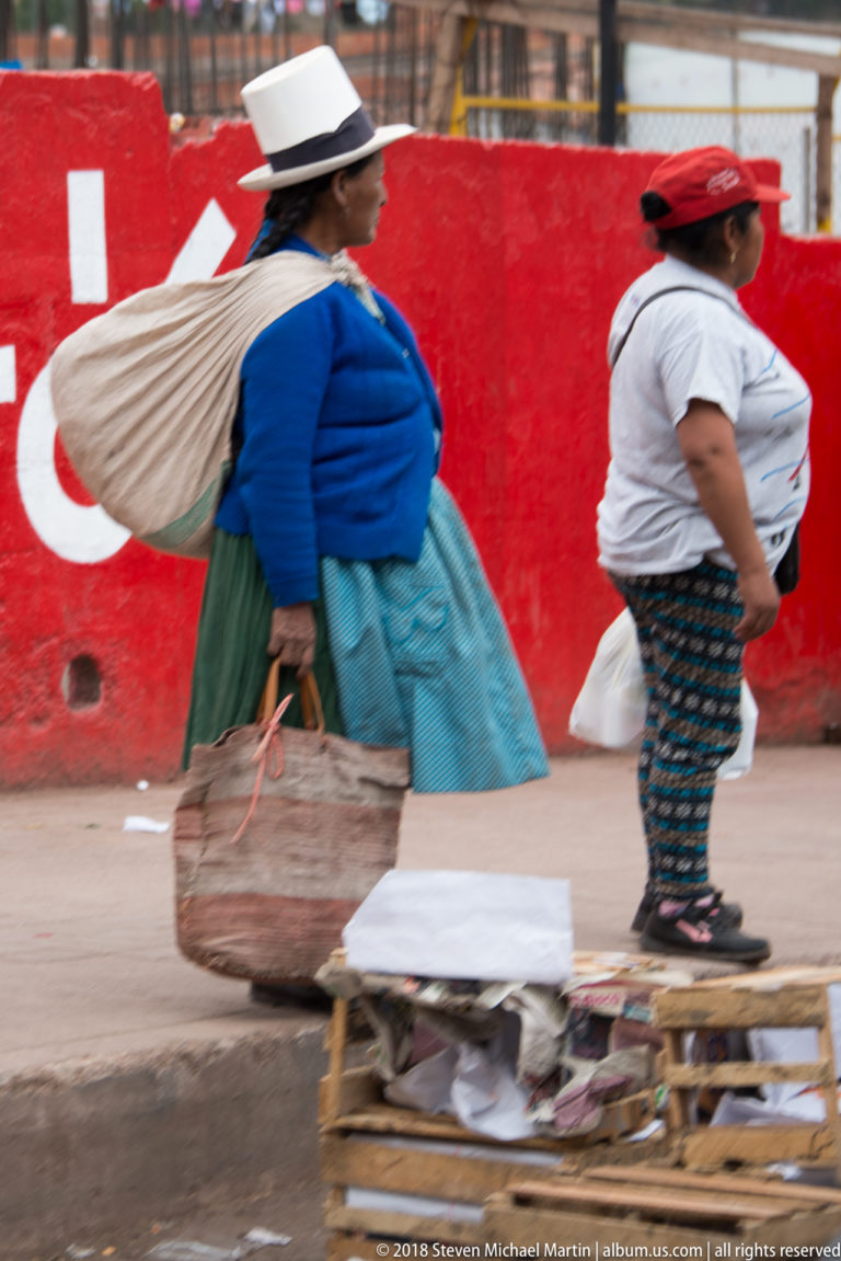 Two women walking past a low street-market display, one in a white hat, blue jacket, and turquoise skirt.