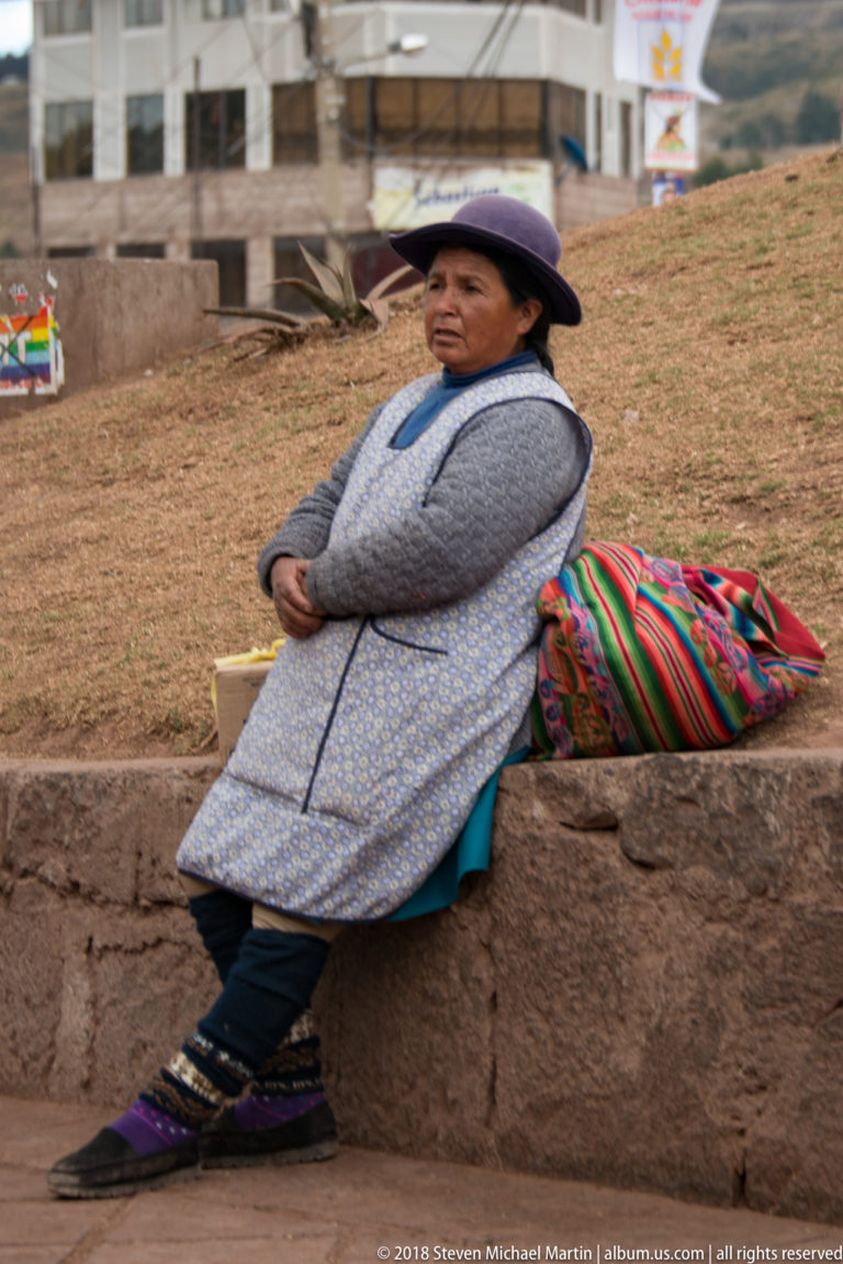 Woman in a purple bowler-style hat seated beside a large brightly striped woven bundle.