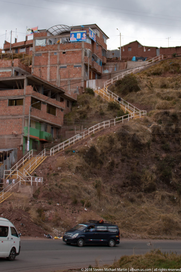 Red-brick buildings and a white-railed switchback stairway rise across a dry hillside above a paved road.