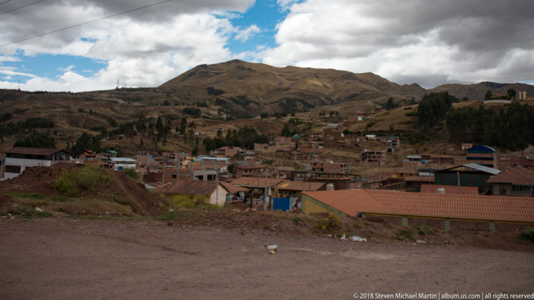 Brick and tile-roof buildings spread across a highland settlement below a dry rounded Andean hillside.
