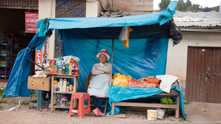 Woman seated beside a blue-tarp street stall with bottles, packaged goods, and red-orange produce.