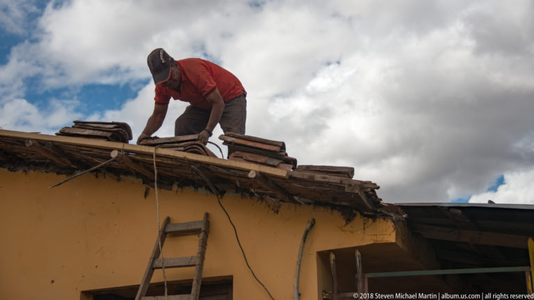 Worker crouching on a sloping clay-tile roof above an ochre wall, with a wooden ladder leaning below.