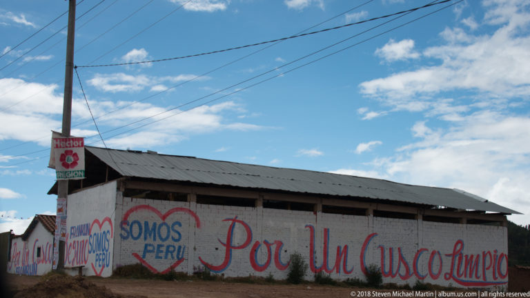 Long white roadside wall painted Somos Perú and Por un Cusco Limpio beneath utility wires and blue sky.