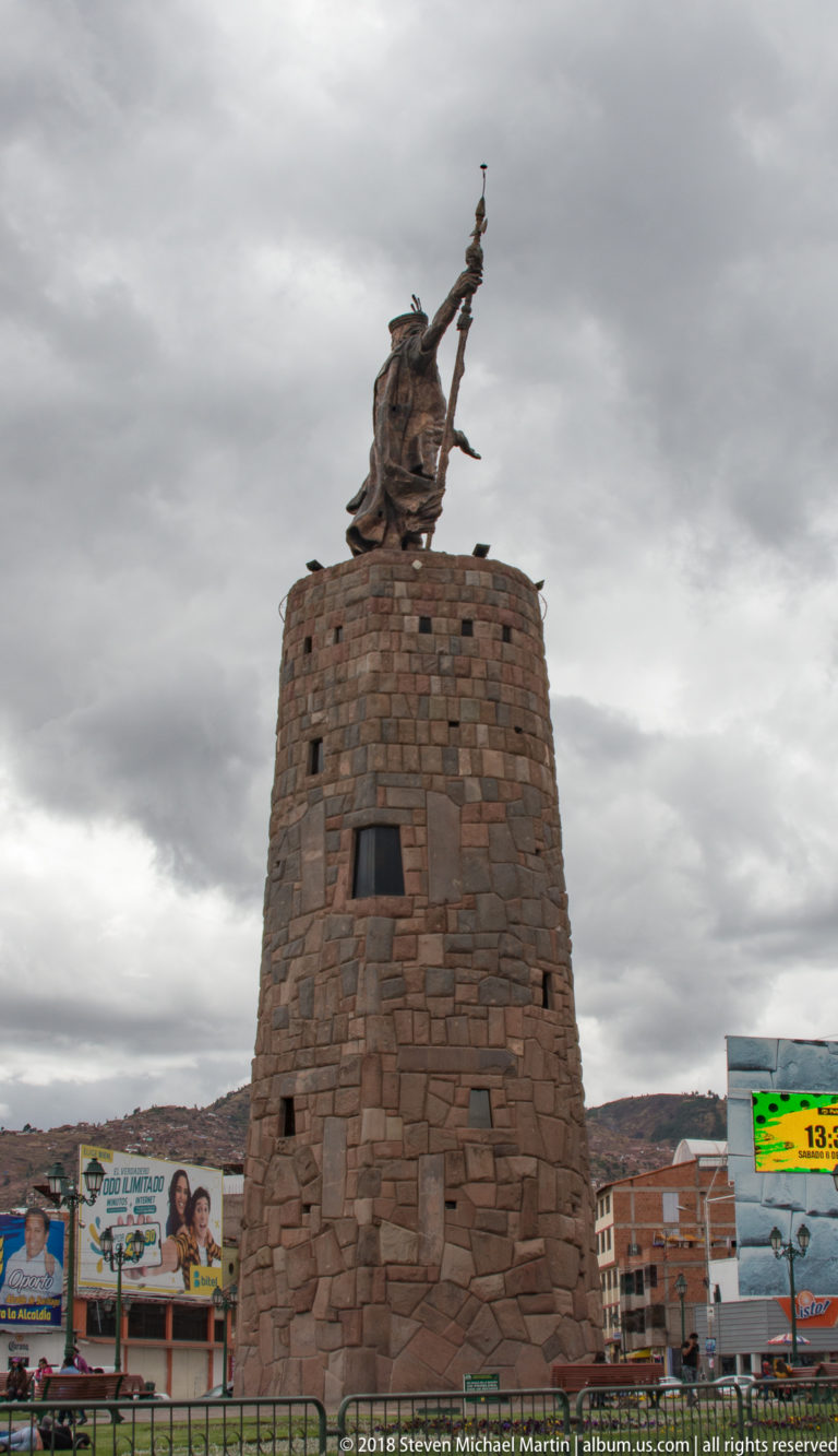 Full view of a tall circular stone monument topped by a warrior figure beneath dark clouds in an urban setting.
