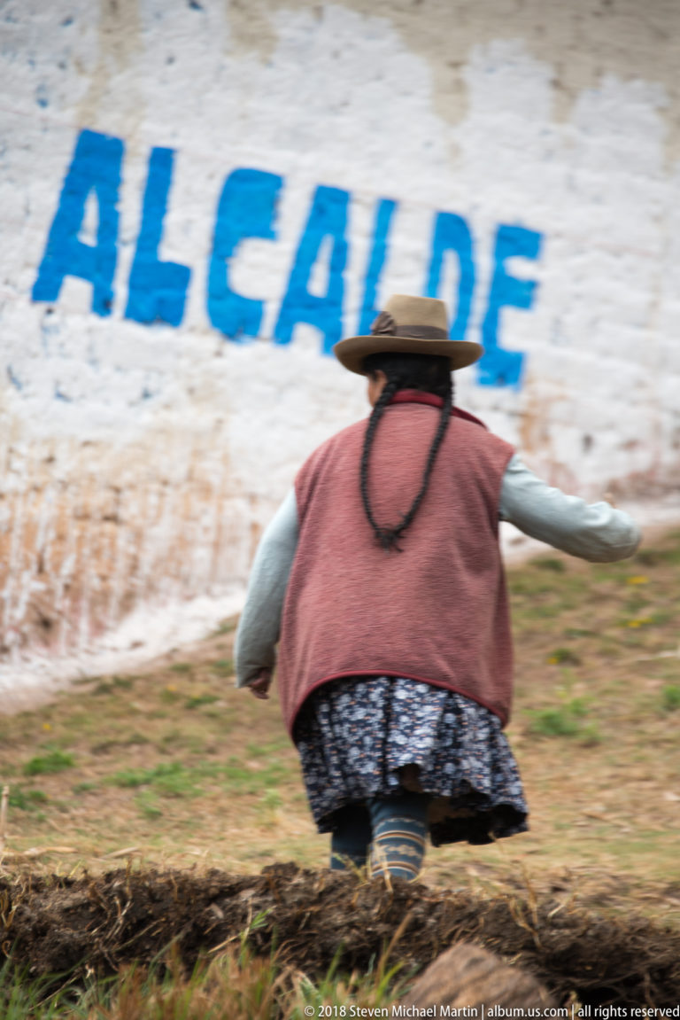 Woman in a broad hat and layered clothing bending over a cultivated strip beside a wall marked ALCALDE.