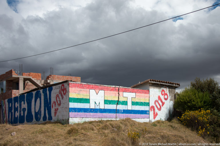 Rainbow-striped roadside political mural with large white MT initials and 2018 markings below dark clouds.