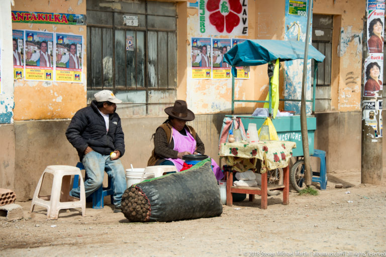 Two seated roadside vendors beside a small cart, stools, buckets, and a large sack of round produce.