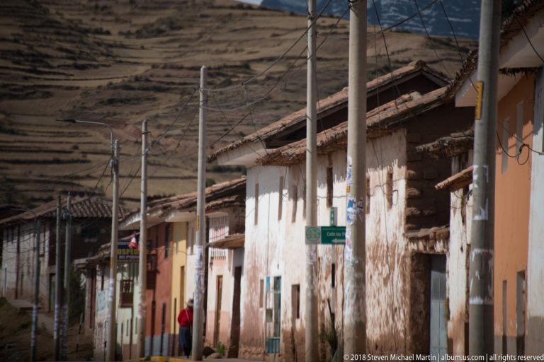 Weathered whitewashed buildings, utility poles, and a pedestrian along a highland street below terraced brown hills.