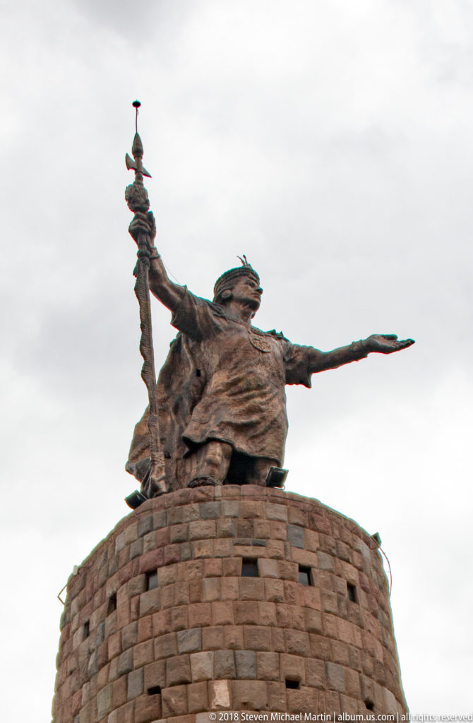Bronze-toned warrior figure extending one arm and raising a staff atop a circular stone tower.