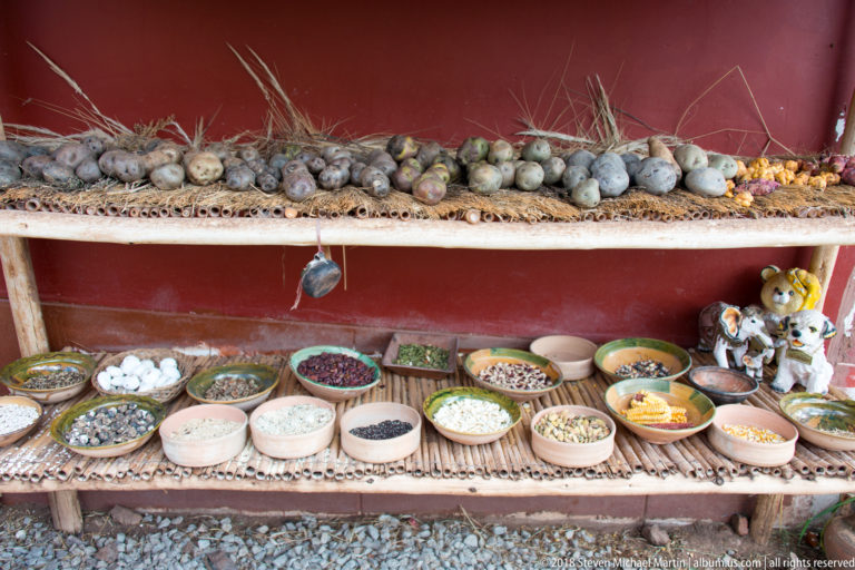 Rustic shelves displaying potatoes and tubers above bowls of assorted grains, seeds, legumes, and dried foods.