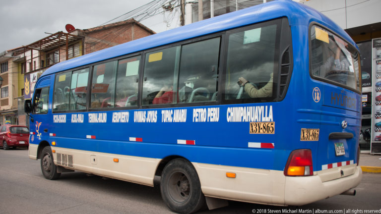 Blue-and-white regional passenger bus with destination names painted along its side, traveling through a city street.