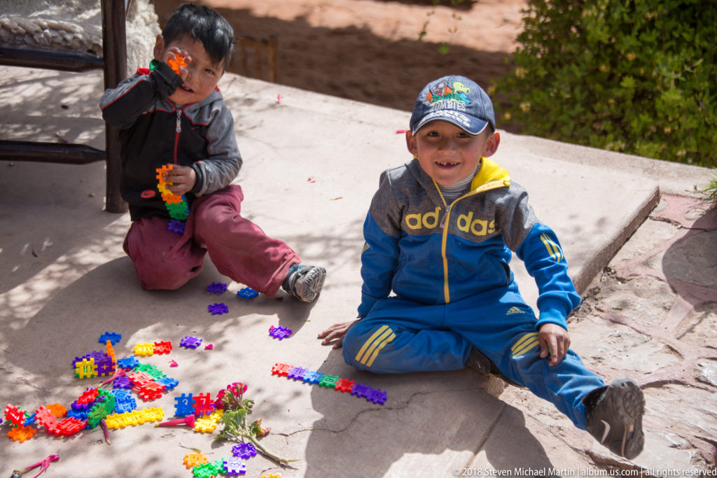 Two children sitting outdoors on paving among scattered brightly colored interlocking plastic construction pieces.