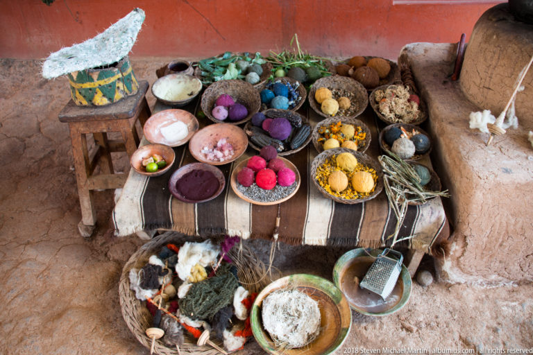 Overhead view of colored yarn, plant materials, fleece, bowls, baskets, vessels, and tools arranged for a textile demonstration.