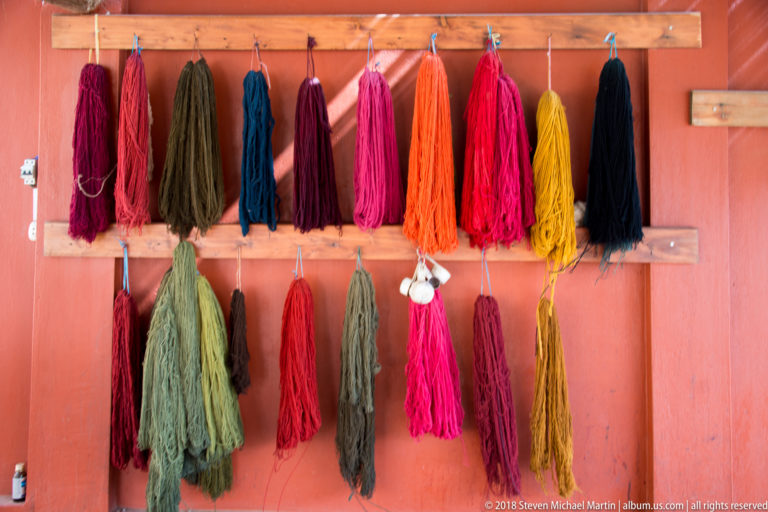 Two rows of long yarn skeins in red, green, blue, orange, pink, yellow, and black hanging on a terracotta wall.