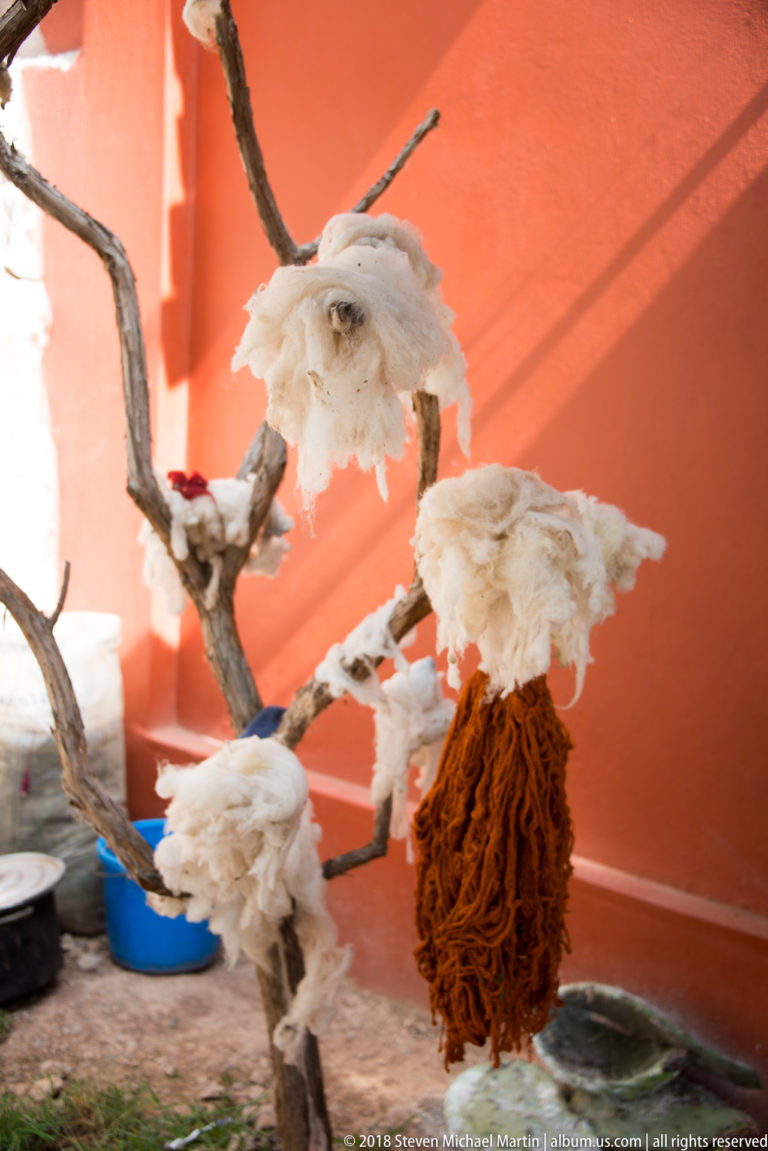 White fiber tufts and a dense orange-brown yarn bundle hanging from bare branches against a terracotta wall.