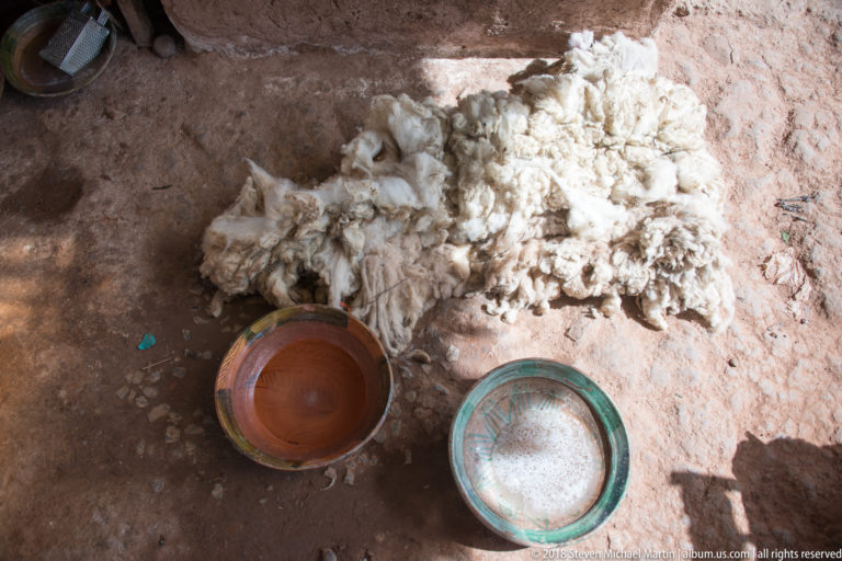 Loose white fleece on an earthen floor beside two shallow ceramic bowls containing brown and pale substances.