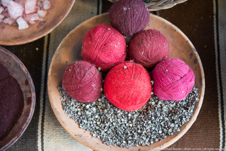 Seven burgundy, red, and pink yarn balls stacked in a shallow bowl above small gray granular material.