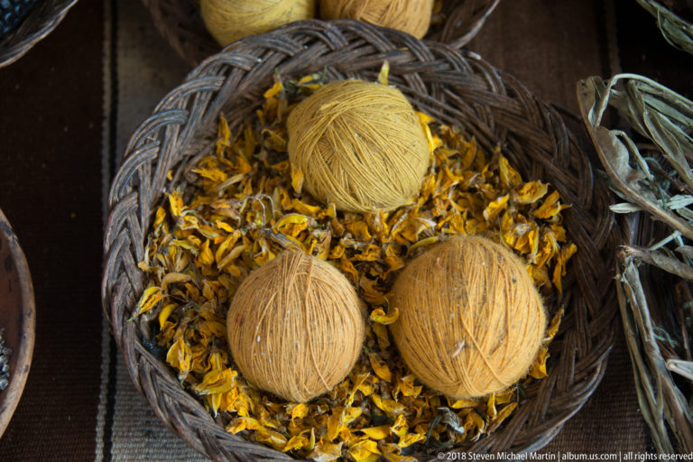Three ochre and mustard yarn balls resting on a dense bed of dried yellow flower petals in a basket.