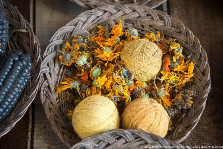 Yellow and ochre yarn balls resting among orange-yellow flower heads and plant fragments in a woven basket.