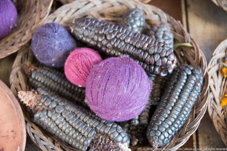 Purple and pink yarn balls arranged among several ears of dark purple-black maize in a woven basket.