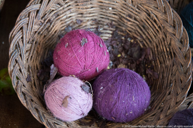 Pale pink, magenta, and purple yarn balls resting over dark reddish material in a woven basket.