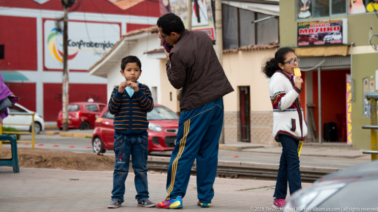 Adult standing with two children eating frozen treats on a sidewalk beside parked cars and storefronts.