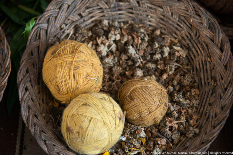Three pale yellow and ochre yarn balls resting on small gray-brown granular material in a woven basket.