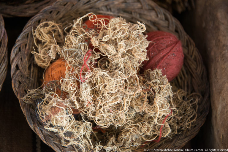 Tangled pale plant-like fibers covering red and orange yarn balls inside a woven basket.