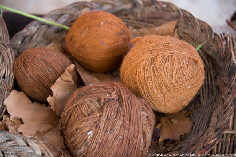 Brown, rust, and orange yarn balls resting among dry bark-like fragments in a woven basket.