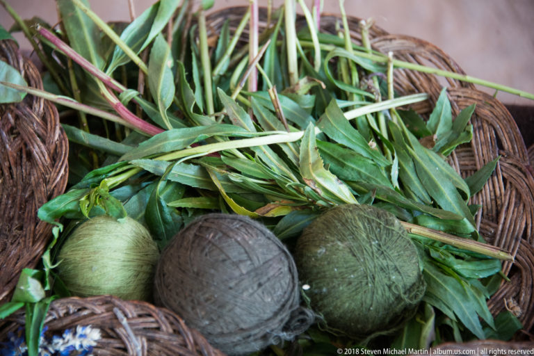 Fresh green leaves and several balls of muted green yarn arranged together in a woven basket.